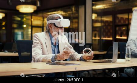 Businessman in VR headset using wireless controllers and laptop, gesturing, watching data in virtual reality. Work in modern office of hi-tech company. Cyberspace digital technology Stock Photo
