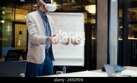 Male engineer using wireless controllers, gesturing, examining blueprint from flipchart in virtual reality, watching data, working in modern office of hi-tech company. Cyberspace digital technology. Stock Photo