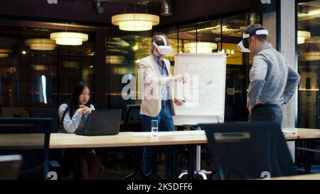 Multi-ethnic engineers in VR headsets, using laptop and wireless controllers, examining blueprint, watching data, working in modern office of hi-tech company. Cyberspace digital technology Stock Photo