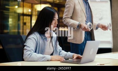 Man using wireless controllers, examining blueprint in virtual reality. Asian female engineer watching data on laptop. Work in modern office of engineering company. Cyberspace digital technology Stock Photo