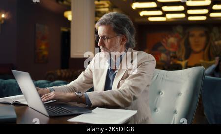 Mature businessman using laptop, working online on startup project, business strategy, analyzing papers and documents in office cafe. Worker of E-commerce marketing company Stock Photo