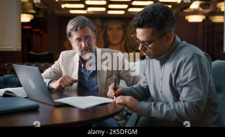 Mature man using laptop, talking with serious businessman, showing papers, documents. Business meeting in office restaurant. Signing contract for new economic strategy of company Stock Photo