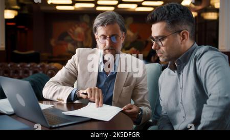 Mature man using laptop, talking with serious businessman, showing papers, documents. Business meeting in office restaurant. Signing contract for new economic strategy of company Stock Photo