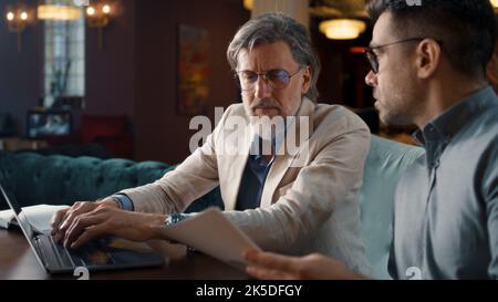 Male colleagues talking, using laptop, working with papers and documents, having business meeting in office restaurant. Teamwork on strategy of E-commerce company Stock Photo