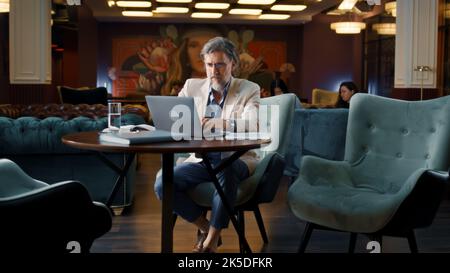 Mature businessman using laptop, working online on startup project, business strategy, analyzing papers and documents in office cafe. Worker of E-commerce marketing company Stock Photo