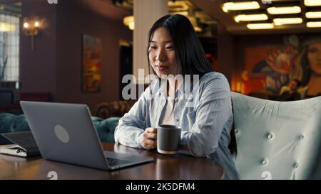 Asian businesswoman looking at screen of laptop, watching business training, working online on startup project, drinking coffee in office cafe. Worker of E-commerce marketing company Stock Photo