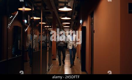 Male coworkers walking down in hallway together, talking about work, discussing startup project or business strategy, using digital tablet computer. Marketing company office corridor Stock Photo
