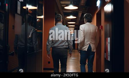 Male colleagues in suits walking down in hallway together, talking about work, discussing startup project or business strategy. Marketing company office corridor Stock Photo