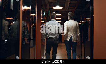 Male colleagues in suits walking down in hallway together, talking about work, discussing startup project or business strategy. Marketing company office corridor Stock Photo