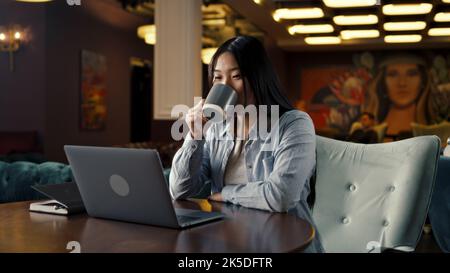 Asian businesswoman looking at screen of laptop, watching business training, working online on startup project, drinking coffee in office cafe. Worker of E-commerce marketing company Stock Photo