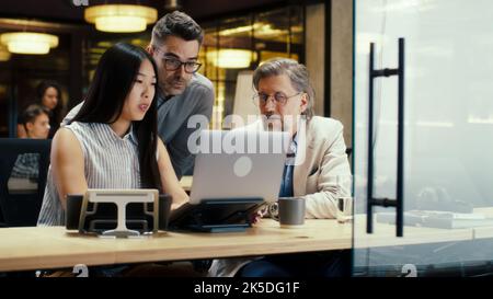 Diverse creative office workers talking and having working conference in modern office, using laptop and digital tablet. Businesspeople working on startup project. Discussion of company strategy Stock Photo