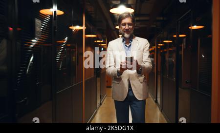 Successful focused businessman, walking in office corridor with phone, surfing the internet, having online conversation. Male worker in hallway of marketing company modern business center Stock Photo