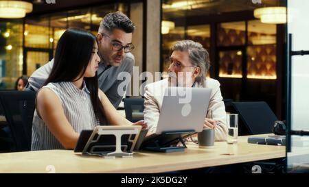 Diverse creative office workers talking and having working conference in modern office, using laptop and digital tablet. Businesspeople working on startup project. Discussion of company strategy Stock Photo