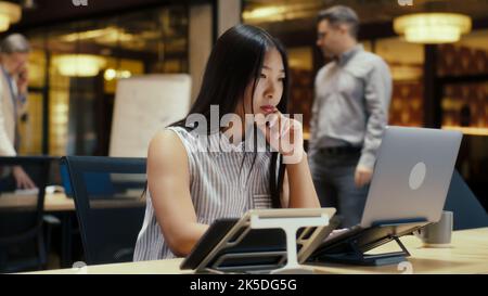 Asian woman working on business project in modern office near coworkers using laptop and smartphone. E-commerce marketing company Stock Photo