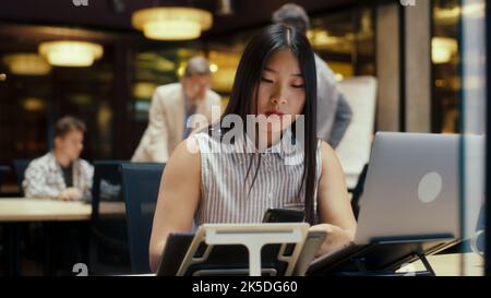 Asian woman working on business project in modern office near coworkers using laptop and smartphone. E-commerce marketing company Stock Photo
