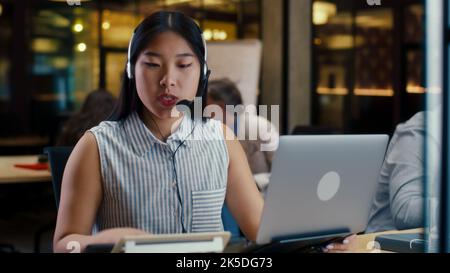 Asian woman in headset talking with client, using laptop and digital tablet. Female operator working in call center office of marketing company Stock Photo