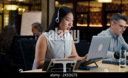 Asian woman in headset talking with client, using laptop and digital tablet. Female operator working in call center office of marketing company Stock Photo