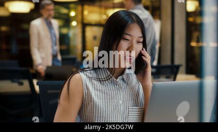 Asian woman talking with client by phone while working in modern office near colleagues, using laptop and digital tablet. Business woman discussing e-commerce startup. Strategy of the company Stock Photo