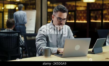 Male office worker, using tablet computer and laptop, typing on keyboard, working in modern office on startup project or business strategy. Programmer developing website in IT company Stock Photo