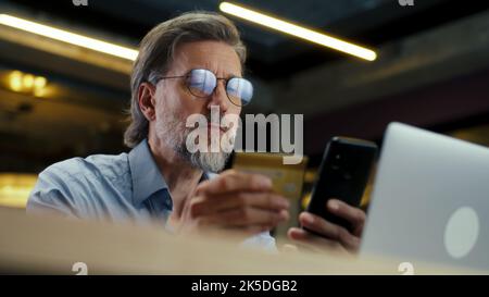 Businessman using phone, transferring money on bank card, paying for service online during work in modern office of marketing company. E-commerce business center Stock Photo