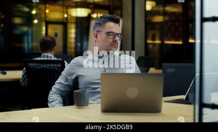 Creative male office worker listening to music, using laptop and digital tablet computer, working in modern office near colleagues on startup project. Work on business strategy of company Stock Photo