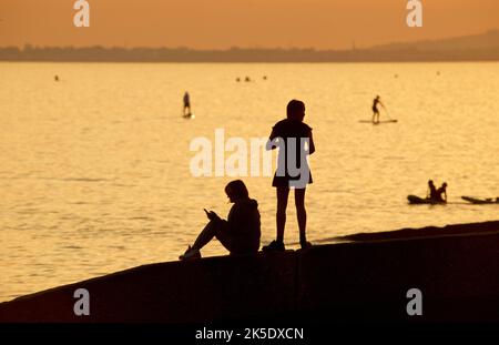 Silhouetted shapes of people sitting and standing on a groyne on a beach, Brighton & Hove. The sky aglow with the fading light of the day. Paddleboarders on the sea in the distance. England, UK Stock Photo
