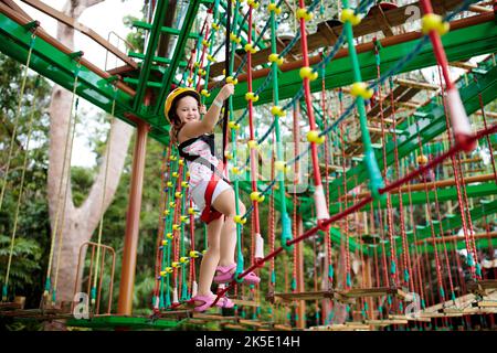 Child in forest adventure park. Kids climb on high rope trail. Agility and climbing outdoor amusement center for children. Stock Photo