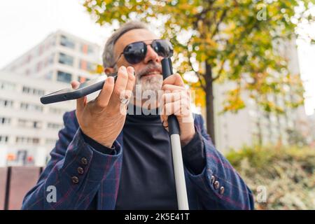 Elderly gray-haired caucasian entrepreneur with low vision in sunglasses sitting on bench, having a phone call using speaker mode, and holding white walking crane with one hand. Outdoor closeup shot. High quality photo Stock Photo