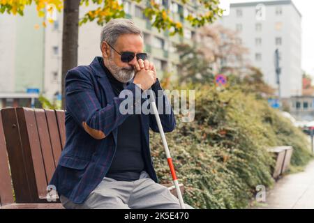 Caucasian gray-haired adult good-looking man in sunglasses with complete loss of sight sitting on bench in park and holding white cane next to his face. Outdoor shot. High quality photo Stock Photo