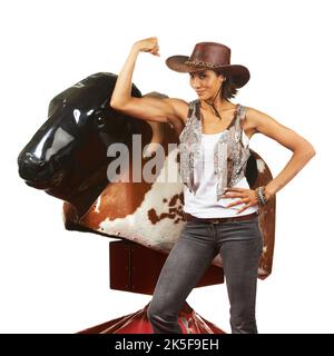 Shes conquered the mechanical bull. Studio shot of a beautiful young cowgirl standing next to a mechanical bull against a white background. Stock Photo
