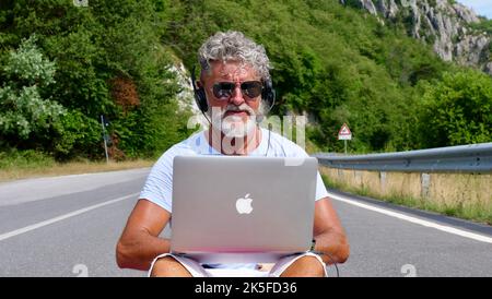 Gray-haired elderly man freelancer with a beard in sunglasses, working on a laptop on nature, road, mountains. A crazy and extraordinary old man sits on a yoga mat in the summer Stock Photo