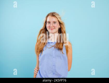 Loose haired teen girl stand with hands keeping behind back on empty blue background. Portrait of smiling school aged child in stylish glasses and Stock Photo