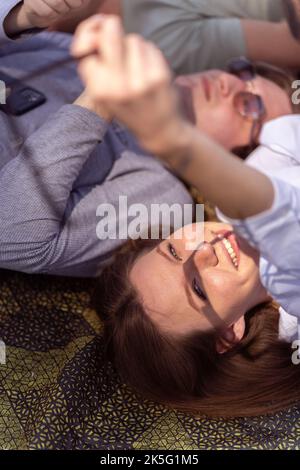 Portrait of young beautiful woman holding sunglasses. Couple relaxing while laying down on green blanket in the park Stock Photo