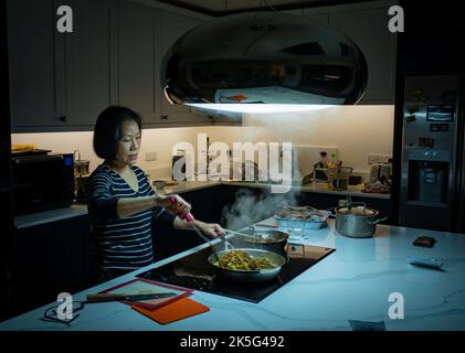 An Asian woman from Vietnam cooks a stir fry meal in her modern kitchen in Billingshurst, West Sussex, UK. Stock Photo