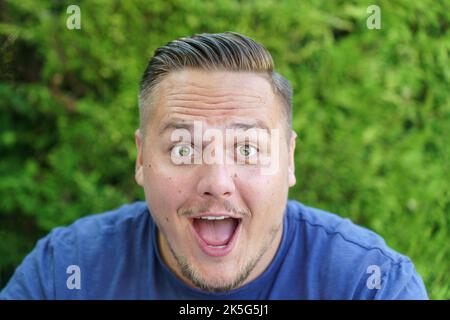 Young man gawping at the camera with mouth open and wide surprised eyes in close up outdoors in the garden Stock Photo
