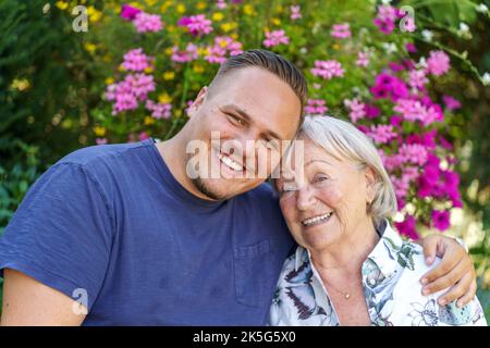 Happy family portrait of a young man with his Grandmother posing arm in arm outdoors in a garden Stock Photo