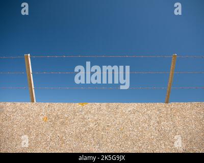 A high wall with three layers of barbed wire on the top to deter intruders. Taken on a sunny day with a clear blue sky. Security concept. Stock Photo
