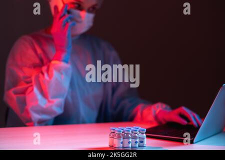 Covid-19 vaccine. Online research. Innovative technology. Female scientist in ppe working on laptop speaking on mobile phone with vial dose bottles in Stock Photo