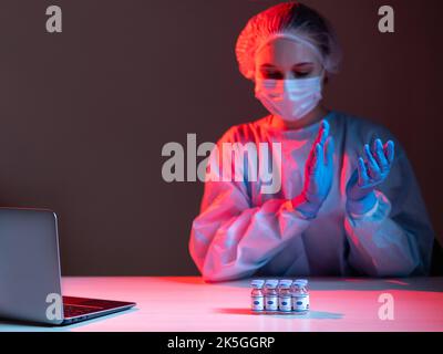 Effective vaccine. Stop covid-19. Approved immunization. Female doctor in ppe face mask gloves applauding to vial dose bottles at workplace with lapto Stock Photo