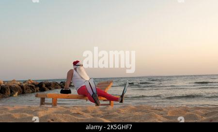 funny santa claus sunbathes. santa, in sunglasses and flippers, is sitting on lounger, on beach by the sea and listening to music, relaxing. santa claus summer vacation, at the seashore. High quality photo Stock Photo