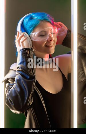 Music joy. Techno playlist. Synth wave. Happy inspired cyberpunk woman in LED neon light listening to tunes in earphones on dark background. Stock Photo