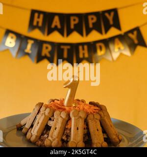 Concept of dog's birthday. Cake for pet made of cookies in shape of meat bones, carrots and golden number of 1 candle. Black flags with inscription ha Stock Photo