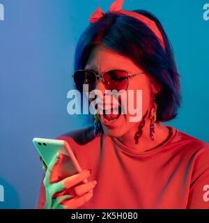Mobile conflict. Phone argue. Communication problem. Angry woman in red neon light complaining on smartphone conversation isolated on blue background. Stock Photo