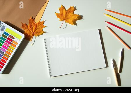 Autumn composition. Watercolor paint and brushes, open blank spirale sketchbook, colored pencils and autumn maple fall leaves on white desk background Stock Photo