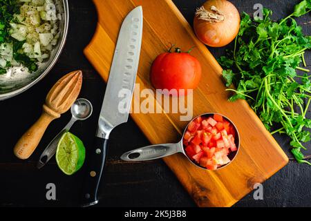 Preparing Raw Ingredients to Make Pico de Gallo: Diced tomatoes in a measuring cup surrounded with other herbs and vegetables Stock Photo