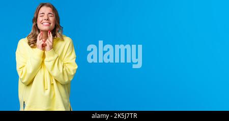 Wishing all well. Hopeful optimistic blond girl in yellow hoodie praying god make wish come true, cross fingers good luck pleading with closed eyes Stock Photo