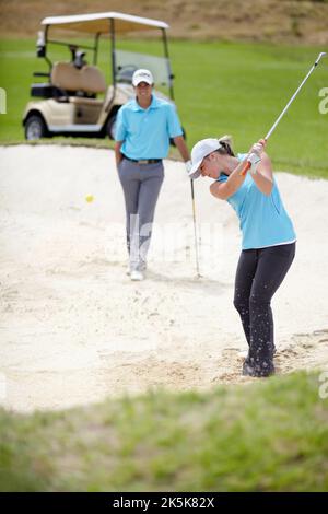 A chip off the old block. A young female golfer chipping her ball out of a bunker while her male partner looks on from behind. Stock Photo