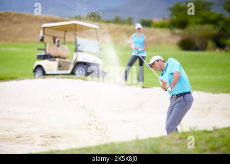 The test of a true golfer. A young male golfer chipping his ball out of a bunker while his female partner looks on from the fairway. Stock Photo