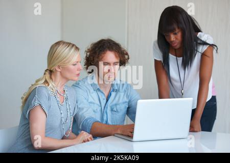 Bringing their ideas to the table. three coworkers gathered around a laptop in the office. Stock Photo