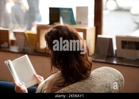 Hanging on every word. A rearview shot of a young woman reading a book by window light. Stock Photo